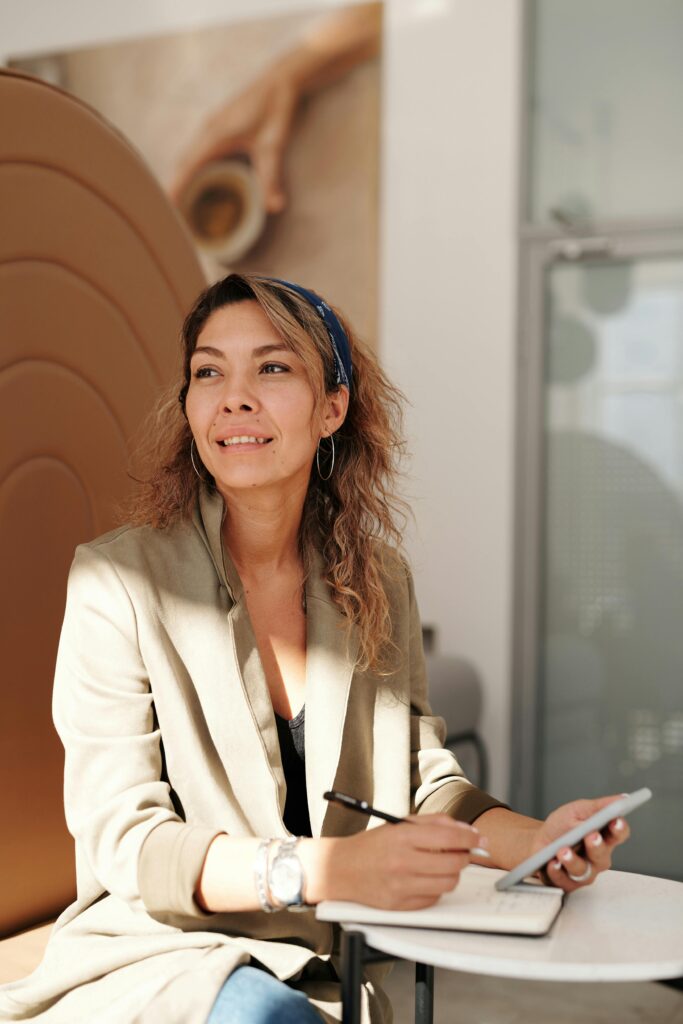 Confident woman in a blazer working on a smartphone and notebook, indoors with sunlight.