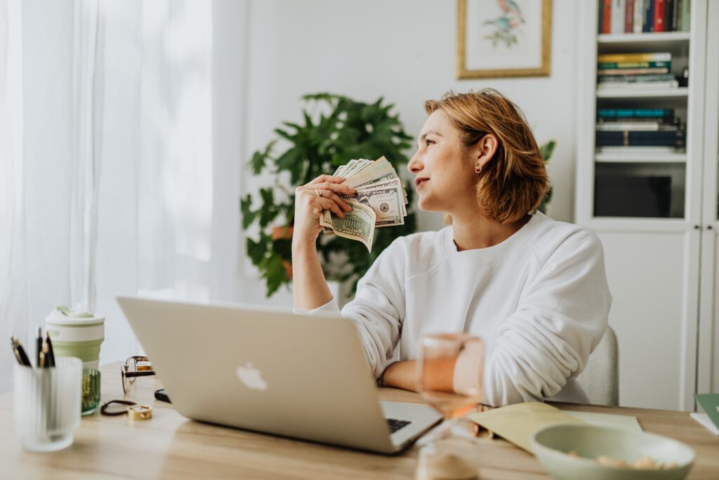 Woman sitting at desk holding cash while working on a laptop in a cozy home office setting.