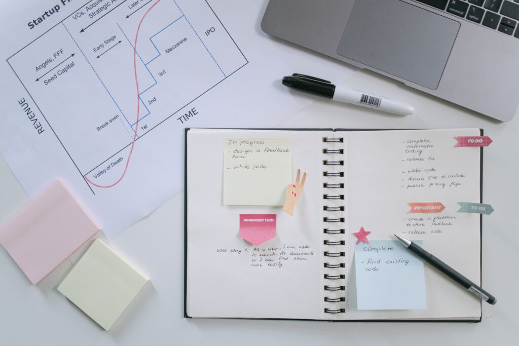 The Difference Maker's Playbook: How Conscious Entrepreneurs Can Grow Their Business and Impact Flat lay of a workspace with an open notebook, sticky notes, planning chart, and laptop for productivity and organization.