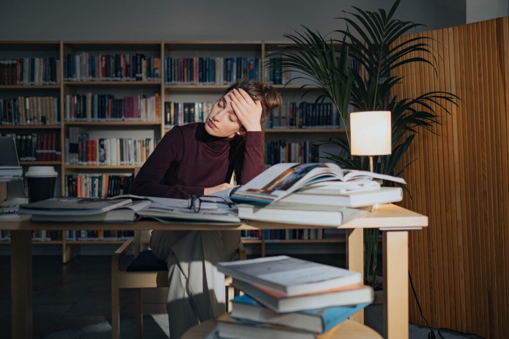 A tired woman surrounded by books, studying in a library, feeling overworked and burnout.